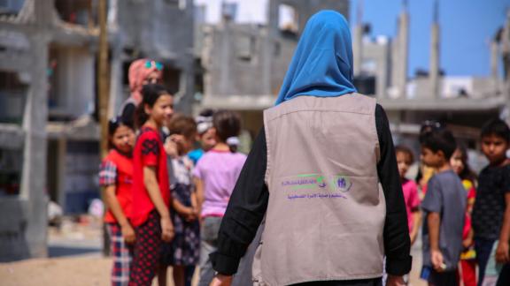 A photo of a person wearing a jacket and a headcover walking towards a group of children, with their back to the camera.